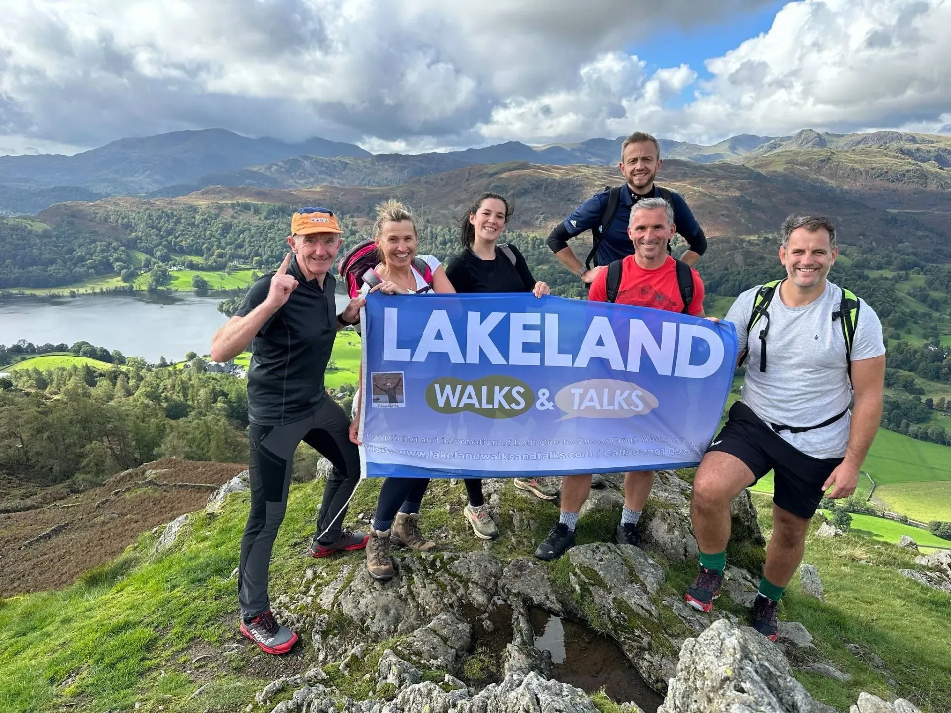 group of people holding a Lakeland Walks & Talks sign on top of a mountain